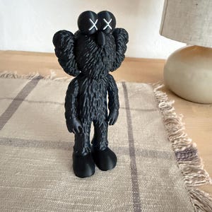 Puede incluir: Una figura negra con una superficie texturizada similar al pelaje y orejas grandes y redondeadas. El personaje presenta dos ojos prominentes marcados con s&iacute;mbolos 'X' blancos, una nariz peque&ntilde;a y pies lisos y redondeados.