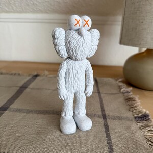 Puede incluir: Una figura KAWS blanca y texturizada con apariencia peluda, que presenta dos ojos redondos marcados con s&iacute;mbolos X naranjas, orejas grandes y pies redondeados. El personaje se encuentra en posici&oacute;n vertical con los brazos ca&iacute;dos.
