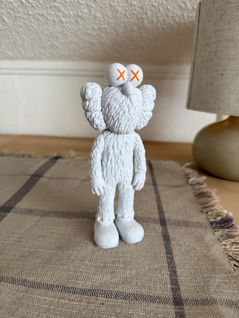 Puede incluir: Una figura KAWS blanca y texturizada con apariencia peluda, que presenta dos ojos redondos marcados con s&iacute;mbolos X naranjas, orejas grandes y pies redondeados. El personaje se encuentra en posici&oacute;n vertical con los brazos ca&iacute;dos.