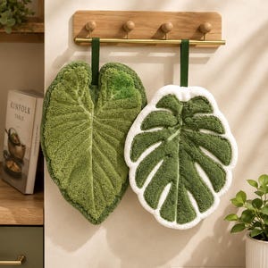 Könnte beinhalten: Zwei blattförmige Hängetücher aus strukturiertem grünem Stoff. Ein Tuch ist wie ein herzförmiges grünes Blatt geformt, das andere zeigt ein dunkelgrünes Monstera-Blattmuster mit einem dicken weißen Rand. Beide hängen an dunkelgrünen Stoffschlaufen.