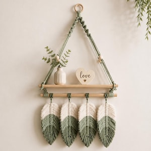 May include: A wall-hanging macrame shelf featuring sage green cord, a wooden plank, and a wooden rod. Four macrame feathers in cream and sage green hang from the rod. A small white vase with greenery and a heart-shaped sign with the word love sit on the shelf.