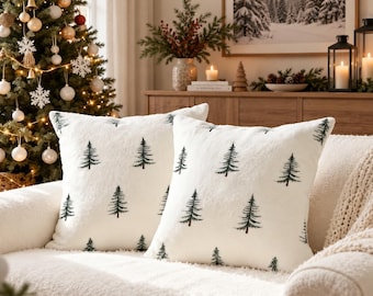 May include: Two square throw pillows with a soft, off-white, textured fabric cover. Each pillow features a repeating pattern of small, dark green pine tree illustrations distributed evenly across the front surface of the cushion.