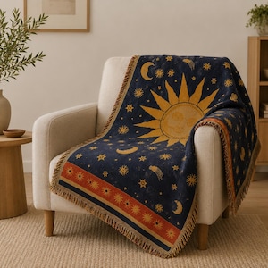 Peut inclure: Un plaid tissé présentant un fond bleu foncé avec un grand soleil doré, des croissants de lune et diverses étoiles. La couverture possède une bordure décorative avec un motif de soleil répétitif en orange et or, finie avec des franges.