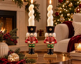 May include: Two nutcracker candle holders featuring red, green, and black uniforms with gold accents. Each nutcracker stands on a gold base and holds a lit, white, spiral-shaped taper candle. The figures have painted faces with black moustaches.