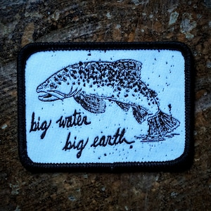 May include: A blue and white embroidered patch with a black outline. The patch features a black and white illustration of a fish with the text "big water big earth".