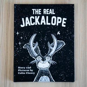 May include: A book cover with a black background and white text that reads "THE REAL JACKALOPE." A cartoon jackalope with antlers is centered below the title. The author's name, "Colin Clancy," is at the bottom.