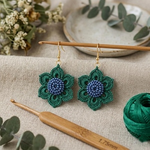 May include: A pair of dangle earrings featuring a six-petaled green crocheted flower design with a central cluster of small blue beads. The earrings are attached to gold-toned metal hooks and are displayed alongside a 1.75 mm bamboo crochet hook.