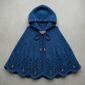 May include: A deep blue knitted hooded cape featuring an intricate lace hem with small silver celestial charms, including stars and moons. The garment has a silver metal clasp at the neck with two braided cords and silver beads along the front edge.
