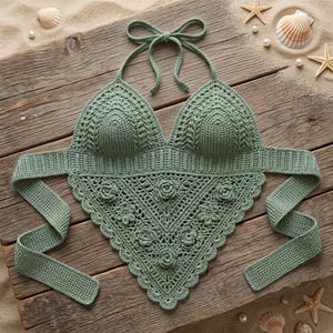 May include: A sage green crochet halter top featuring a triangular bodice with a scalloped edge, intricate floral motifs, and long ties at the neck and back. The garment is crafted with a detailed stitch pattern and has a soft, textured appearance.