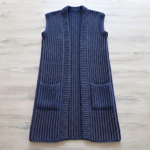 May include: A sleeveless, knee-length cardigan vest in a deep navy blue color. This garment features a prominent vertical ribbed knit texture throughout, a wide ribbed collar border, and two large, square patch pockets on the front lower section.