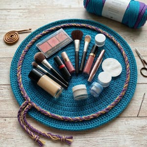 Puede incluir: Una alfombrilla redonda de ganchillo color verde azulado con un borde trenzado en hilo morado y dorado. La alfombrilla está dispuesta con varios artículos de maquillaje, incluyendo brochas, una paleta de colorete, pintalabios, rímel y tarros.
