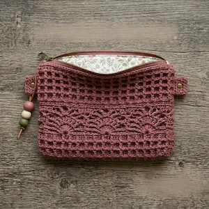 Puede incluir: Un bolso de ganchillo en color rosa empolvado con un patrón de rejilla y una banda decorativa de punto de abanico. El bolso tiene una cremallera dorada, un forro de tela con estampado botánico y un tirador de cuero con tres cuentas de madera.
