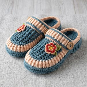 Könnte beinhalten: Ein Paar handgehäkelte Clogs in einem tiefblauen und sanften pfirsichfarbenen Design. Jeder Schuh verfügt über ein strukturiertes Gittermuster auf dem Obermaterial, einen gerippten pfirsichfarbenen Rand und eine kleine, bunte Blumenapplikation mit grünen Blättern.