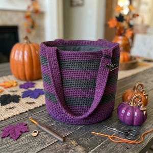 May include: A cylindrical crochet tote bag featuring horizontal stripes in alternating shades of dark purple and charcoal gray. The bag has two matching crochet handles and a small black metal bat-shaped charm attached to one of the straps.