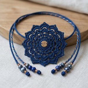 Könnte beinhalten: Eine dunkelblaue gehäkelte Mandala-Halskette mit einem zentralen Metallanhänger, der ein kleines Pflanzensymbol zeigt. Das Schmuckstück besteht aus filigraner Fadenarbeit, kleinen Metallperlen und Enden, die mit dunkelblauen Perlen und Metallakzenten verziert sind.