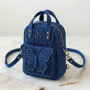 May include: A dark blue crochet backpack featuring a prominent butterfly design on the front pocket, a gold-toned zipper closure, two top handles, and adjustable shoulder straps. The textured yarn construction includes intricate cable knit patterns.