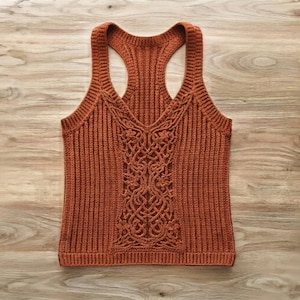 May include: A rust-colored sleeveless tank top featuring a racerback design and a ribbed knit texture. The front panel displays an intricate, raised cable knit pattern with floral motifs integrated into the center, creating a textured look.