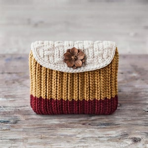 May include: A small rectangular pouch featuring a cream-colored crocheted flap with a basket-weave pattern, secured by a dark brown wooden flower button. The body of the pouch is knitted with a ribbed texture in mustard yellow and deep burgundy.