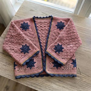 May include: A dusty rose crochet cardigan featuring a repeating openwork diamond pattern and dark navy blue star-shaped motifs. The garment is finished with a scalloped dark navy blue border and a thin mustard yellow accent line along the edges.