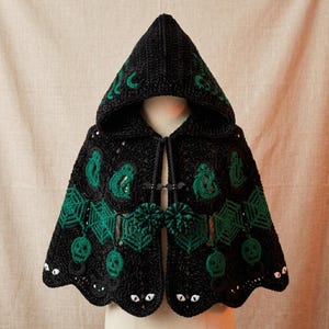 May include: A black hooded crochet cape featuring green motifs of ghosts, spiderwebs, and skulls. The garment includes small black bat-shaped toggles at the neck, two dark green pom-poms, and a scalloped hem decorated with white cat eye details.
