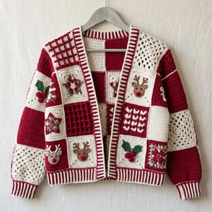 Può includere: Un cardigan all'uncinetto a maniche lunghe con un motivo a scacchi composto da quadrati rosso scuro e crema. Il capo è decorato con varie applicazioni, tra cui volti di renna marroni con nasi rossi, foglie di agrifoglio verdi e stelle.