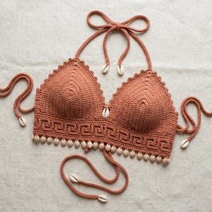 May include: A terracotta-colored crochet bikini top featuring a triangular cup design with a Greek key pattern along the bottom edge. The garment is accented with small white cowrie shells along the hem and ties, with additional shells at the strap ends.