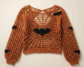 Può includere: Un maglione all'uncinetto a maniche lunghe color arancione bruciato con un motivo aperto a ragnatela su tutto il corpo e le maniche. Il capo è decorato con diverse piccole applicazioni di pipistrelli neri realizzate all'uncinetto.