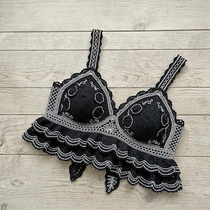 Op de afbeelding: Een zwarte gehaakte crop top met witte geborduurde bloemendetails en geometrische patronen. Het kledingstuk heeft twee brede schouderbandjes, een gestructureerd lijfje met geïntegreerde cups en een dubbellaagse ruchezoom met geschulpte randen.