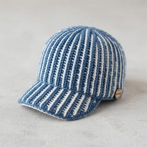 Puede incluir: Una gorra de béisbol de punto con un patrón de rayas verticales en azul y blanco. La gorra tiene una corona redondeada y una visera rígida, con un pequeño detalle de botón de madera de color marrón claro circular en el lateral.