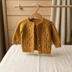 Può includere: Un cardigan a maniche lunghe color giallo senape realizzato in filato lavorato all'uncinetto. Il capo presenta un motivo verticale a trama di foglie su entrambi i pannelli frontali, scollo rotondo, polsini e orlo a coste e cinque bottoni marrone chiaro.
