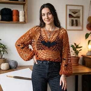 May include: A burnt orange crochet sweater featuring a spiderweb pattern throughout, long sleeves, and a black bat applique centered on the chest. The garment is worn over a black camisole and paired with dark denim jeans, creating a seasonal look.