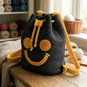 Könnte beinhalten: Ein dunkelgrauer Häkel-Rucksack mit Kordelzug und einem leuchtend gelben Smiley-Gesicht auf der Vorderseite. Die Tasche verfügt über zwei gelbe Schultergurte, einen gelben Kordelzug, zwei kreisförmige gelbe Augen und einen geschwungenen gelben Mund.