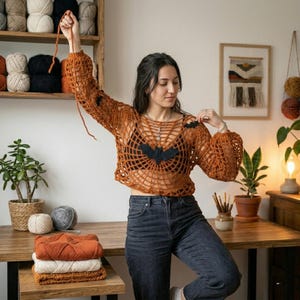 May include: A burnt orange crochet sweater featuring a spiderweb pattern and black bat appliques on the chest and shoulder. The garment has long sleeves with gathered cuffs and a wide neckline, worn over a black top with dark gray denim jeans.