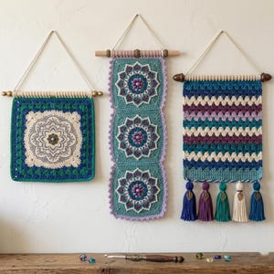 May include: Three wall hangings featuring intricate crochet work. The left piece is a square mandala in cream, blue, and green. The center piece is a vertical strip of three floral mandalas in teal and purple. The right piece has horizontal stripes with five tassels featuring metal leaf charms.