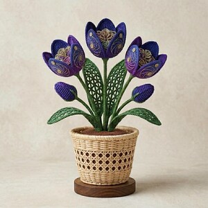 May include: A decorative arrangement of three large purple crocheted tulip-like flowers and two smaller buds, featuring intricate gold thread detailing. The flowers have green crocheted stems and leaves, all housed in a light tan woven wicker basket.