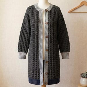 May include: A long-sleeved, knee-length cardigan featuring a dark charcoal gray waffle-knit texture. The garment includes a light gray ribbed collar, cuffs, and front button placket with brown buttons, finished with a solid dark navy blue hem.