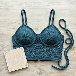 May include: A dark teal crochet crop top featuring molded cups, thin shoulder straps, and a geometric lattice pattern on the bodice. The garment includes long side ties for fastening and is displayed alongside a square wooden plaque with a mandala design.