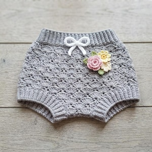 Pode incluir: Um tapa-fraldas de crochê cinza com um padrão de ponto concha texturizado, cós canelado e um pequeno laço de fio branco. Um grupo de três pequenas flores de crochê nas cores rosa, amarelo e creme com folhas verdes está fixado na frente.