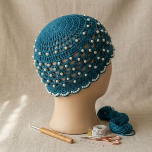 May include: A teal crochet beanie featuring an openwork lattice pattern embellished with numerous small white faux pearls. The hat has a scalloped edge and is displayed on a mannequin head alongside a wooden crochet hook and a measuring tape.