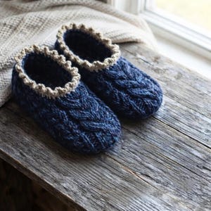 Può includere: Un paio di pantofole lavorate a maglia blu scuro con un motivo a trecce spesso sull'esterno e un bordo smerlato beige chiaro attorno all'apertura. Le pantofole sono realizzate con filato testurizzato e hanno una punta arrotondata.