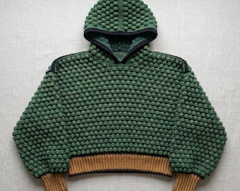 May include: A forest green crochet hoodie featuring a dense bobble stitch texture throughout the body and sleeves. The garment includes a dark navy blue trim along the hood edge and shoulder seams, with a solid tan ribbed hem and sleeve cuffs.