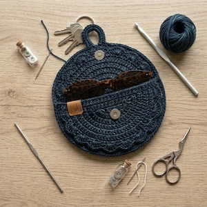 May include: A round, dark blue crocheted pouch with a scalloped edge, a magnetic snap closure, and a small tan leather tag. The pouch holds brown tortoiseshell glasses. Surrounding items include a ball of yarn, crochet hooks, scissors, and keys.
