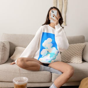 May include: A white long-sleeve crewneck sweatshirt featuring a rectangular graphic print on the front. The graphic depicts a bright blue sky filled with stylized, fluffy white and light blue clouds with soft, rounded edges and subtle gradients.
