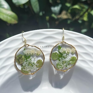 Puede incluir: Un par de pendientes colgantes redondos con marcos de resina transparente y bordes dorados. Cada pendiente contiene flores blancas prensadas y hojas verdes incrustadas en el material transparente, sujetos por ganchos de metal dorado.
