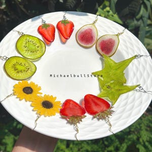 May include: A collection of fruit and flower shaped earrings arranged on a white plate, featuring strawberries, kiwi slices, fig slices, starfruit, and sunflowers. The text MichaelbullStore is printed in the center of the white plate.