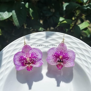 May include: A pair of drop earrings featuring realistic purple and white orchid flower petals with speckled patterns. Each orchid is attached to a thin gold-toned metal hook. The text MichaelbullStore is printed in the centre between the earrings.