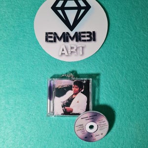 Könnte beinhalten: Eine Miniatur-Nachbildung des Michael Jackson Thriller-Albums, bestehend aus einer transparenten Kunststoffhülle mit Cover-Art und einer kleinen runden Disc. Darüber befindet sich ein rundes weißes Logo mit schwarzem Diamant und dem Text EMMEBI ART.