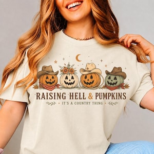 May include: A cream-colored t-shirt featuring a graphic of four jack-o'-lanterns wearing cowboy hats. The text reads "RAISING HELL & PUMPKINS" and "IT'S A COUNTRY THING" with small stars and a crescent moon above the pumpkins.