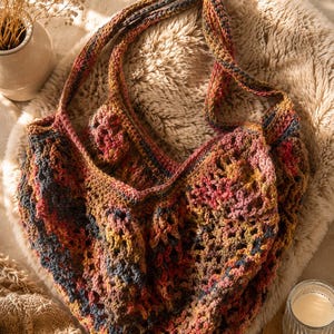 Könnte beinhalten: Eine handgefertigte Häkel-Tragetasche mit einem offenen Netzmuster. Die Tasche ist aus meliertem Garn in einer Mischung aus Erdtönen wie Rost, Senfgelb, Dunkelblau und gedämpftem Rosa gefertigt und verfügt über zwei integrierte Schulterriemen.