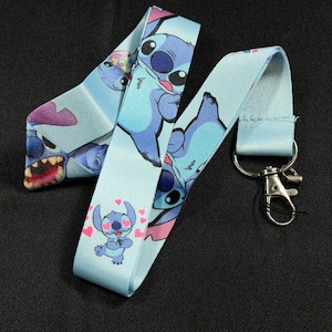 Puede incluir: Un cordón de tela azul claro con un patrón repetitivo del personaje alienígena azul Stitch de Lilo y Stitch. El diseño incluye a Stitch en varias poses, algunas con corazones rosas, y un cierre de mosquetón de metal en tono plateado.